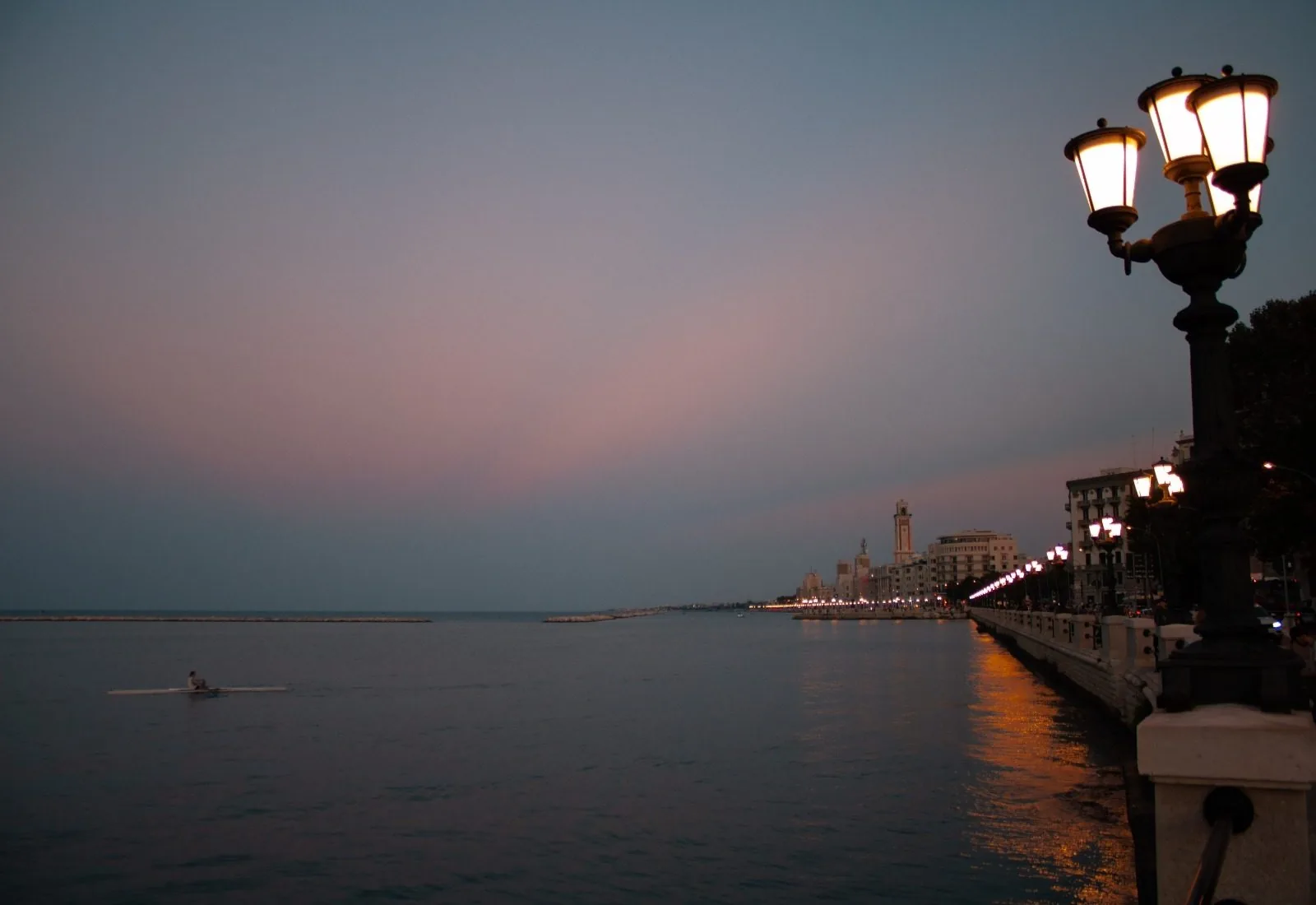 Il lungomare Nazario Sauro di Bari al crepuscolo, con i lampioni accesi riflessi sul mare calmo e una canoa in lontananza